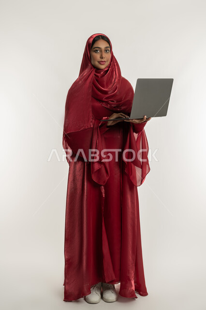 إنجاز المهام والعمل عن بعد, استخدام الأجهزة التقنية الحديثة, بورتريه لامرأة عربية خليجية إماراتية محجبة ترتدي العباءة الحمراء تعمل على جهاز الحاسوب المحمول, شابة سعودية تنظر إلى الكاميرا بإيماءات السعادة والسرور, الجسم بالطول الكامل, خلفية بيضاء