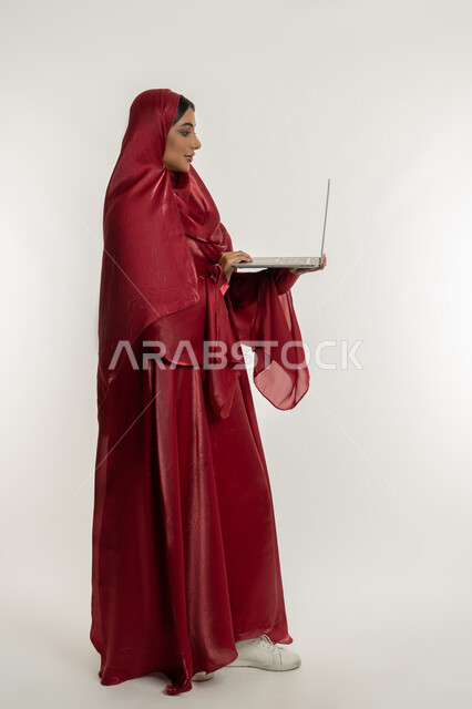 Remote work concept, use of modern and advanced technical devices, side portrait of a veiled Emirati Gulf Arab woman wearing a red abaya working on a laptop, a young Saudi woman completing tasks, full body, white background
