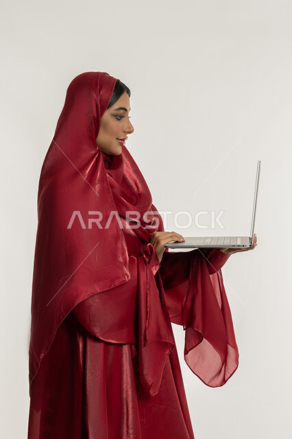 Using modern and advanced technical devices, the concept of remote work, a side portrait of a veiled Emirati Gulf Arab woman wearing a red abaya working on a laptop, a young Saudi woman completing tasks, white background