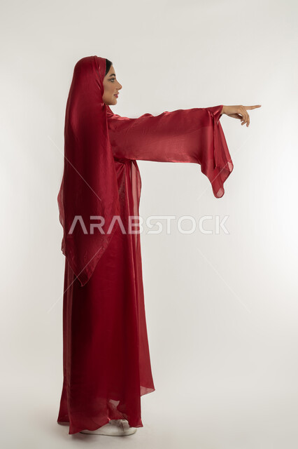 تحديد موقع شيء معين, التوضيح والدلالة لمكان محدد, بورتريه من الجانب لامرأة عربية خليجية إماراتية محجبة ترتدي العباءة الحمراء تشير بإصبع السبابة للأمام، شابة سعودية تقوم بالإشارة إلى عنصر ما, الجسم بالطول الكامل, خلفية بيضاء
