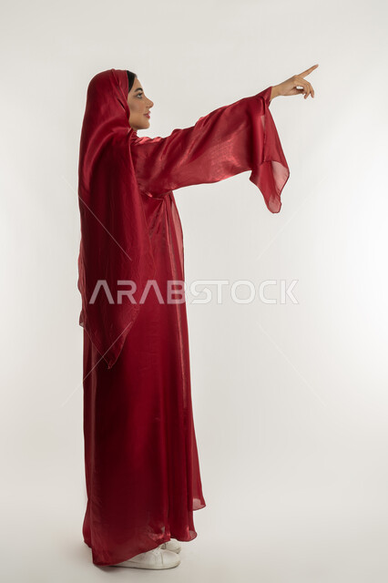 الإشارة إلى عنصر ما, الدلالة والتوضيح لمكان محدد, بورتريه من الجانب لامرأة عربية خليجية إماراتية محجبة ترتدي العباءة الحمراء تشير بإصبع السبابة للأعلى، شابة سعودية تقوم بتحديد موقع شيء معين, الجسم بالطول الكامل, خلفية بيضاء