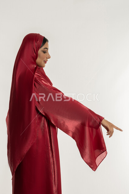 الإشارة إلى عنصر ما, التوضيح والدلالة لمكان محدد, بورتريه من الجانب لامرأة عربية خليجية إماراتية محجبة ترتدي العباءة الحمراء تشير بإصبع السبابة للأسفل، شابة سعودية تقوم بتحديد موقع شيء معين, خلفية بيضاء