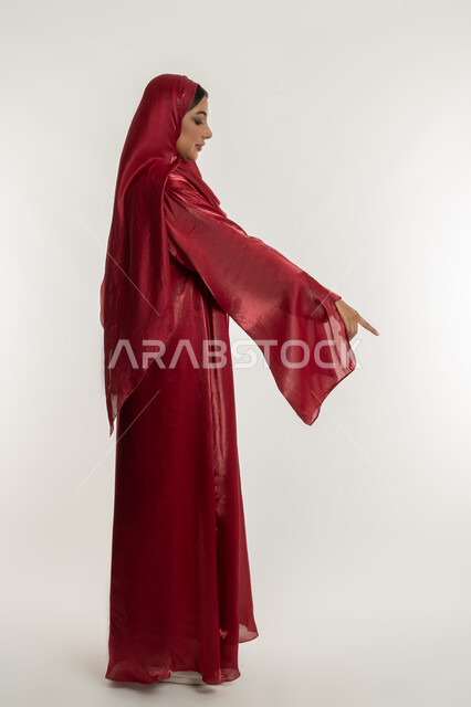 التوضيح والدلالة لمكان محدد, الإشارة إلى عنصر ما, بورتريه من الجانب لامرأة عربية خليجية إماراتية محجبة ترتدي العباءة الحمراء تشير بإصبع السبابة للأسفل، شابة سعودية تقوم بتحديد موقع شيء معين, الجسم بالطول الكامل, خلفية بيضاء