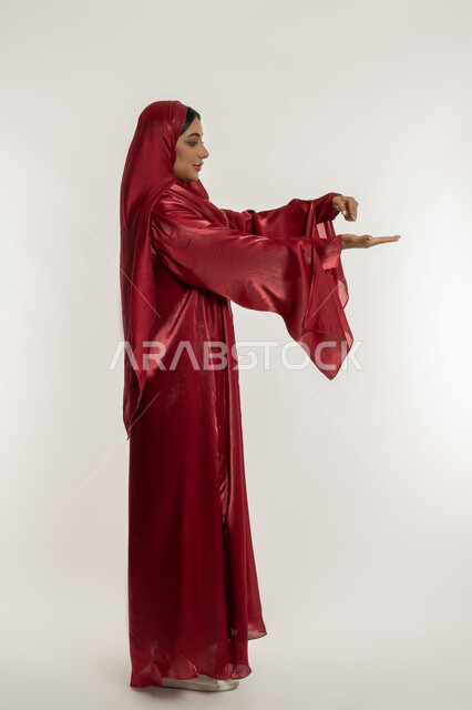 حمل و إمساك عنصر ما, تعابير رفع شيء معين, بورتريه من الجانب لامرأة عربية خليجية إماراتية محجبة ترتدي العباءة الحمراء تمسك بعناصر محددة, شابة سعودية تقف باستقامة واعتدال, الجسم بالطول الكامل, خلفية بيضاء