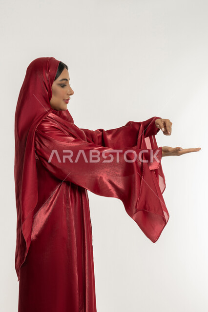 تعابير رفع شيء معين, حمل و إمساك عنصر ما, بورتريه من الجانب لامرأة عربية خليجية إماراتية محجبة ترتدي العباءة الحمراء تمسك بعناصر محددة, شابة سعودية تقف باستقامة واعتدال, خلفية بيضاء