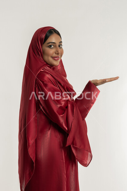 رفع شيء معين, تعابير حمل و إمساك عنصر محدد, بورتريه من الجانب لامرأة عربية خليجية إماراتية محجبة ترتدي العباءة الحمراء ترفع كفها للأعلى, شابة سعودية تنظر إلى الكاميرا بإيماءات السعادة والسرور, خلفية بيضاء