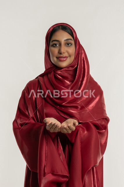 الاجتهاد في الطاعات والأدعية والأذكار, مفهوم العبادة والتقرب إلى الله, بورتريه لامرأة عربية خليجية إماراتية محجبة ترتدي العباءة الحمراء ترفع يديها للأعلى بتعابير الدعاء, شابة سعودية تنظر إلى الكاميرا بإيماءات السعادة والسرور, خلفية بيضاء