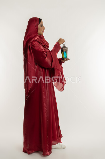 البهجة بقدوم شهر الخير, الاحتفال بالمناسبات الدينية الإسلامية, بورتريه من الجانب لامرأة عربية خليجية إماراتية محجبة ترتدي العباءة الحمراء تمسك بيدها فانوس معدني مزخرف, شابة سعودية تقوم بالتحضير لاستقبال رمضان المبارك, الجسم بالطول الكامل, خلفية بيضاء