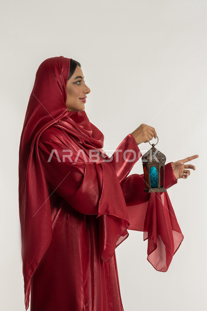 البهجة بقدوم شهر الخير, الاحتفال بالمناسبات الدينية الإسلامية, التحضير لاستقبال رمضان المبارك, بورتريه من الجانب لامرأة عربية خليجية إماراتية محجبة ترتدي العباءة الحمراء تمسك بيدها فانوس معدني مزخرف, شابة سعودية تشير بإصبع السبابة إلى شيء ما, خلفية بيضاء