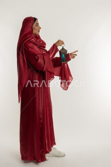 التحضير لاستقبال رمضان المبارك, الاحتفال بالمناسبات الدينية، البهجة بقدوم شهر الخير, بورتريه من الجانب لامرأة عربية خليجية إماراتية محجبة ترتدي العباءة الحمراء تمسك بيدها فانوس معدني, شابة سعودية تشير بإصبع السبابة إلى شيء ما, الجسم بالطول الكامل, خلفية بيضاء