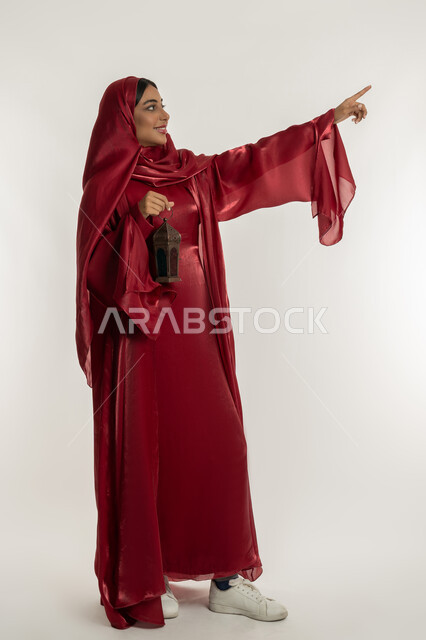 الاحتفال بالمناسبات الدينية الإسلامية، البهجة بقدوم شهر الخير, التحضير لاستقبال رمضان المبارك, بورتريه لامرأة عربية خليجية إماراتية محجبة ترتدي العباءة الحمراء تمسك بيدها فانوس معدني, شابة سعودية تشير بإصبع السبابة إلى شيء ما, الجسم بالطول الكامل, خلفية بيضاء