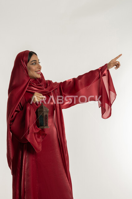 البهجة بقدوم شهر الخير, الاحتفال بالمناسبات الدينية الإسلامية, التحضير لاستقبال رمضان المبارك, بورتريه لامرأة عربية خليجية إماراتية محجبة ترتدي العباءة الحمراء تمسك بيدها فانوس معدني مزخرف, شابة سعودية تشير بإصبع السبابة إلى شيء ما, خلفية بيضاء