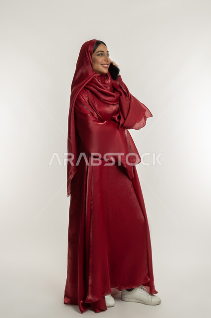 Using modern technology devices, communicating with family and friends, sharing good news and conversations, portrait of a veiled Emirati Gulf Arab woman wearing a red abaya holding a mobile phone in her hand, a young Saudi woman making a phone call, full body, white background