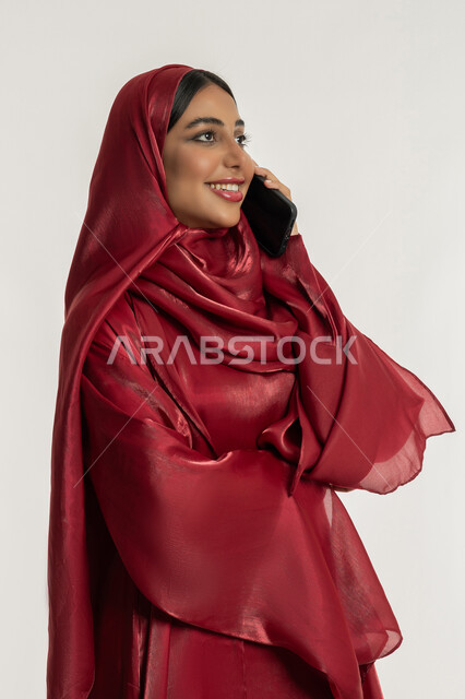 مشاركة الأخبار والأحاديث الجيدة, التواصل مع العائلة والأصدقاء, استخدام الأجهزة التقنية الحديثة في الحياة اليومية, بورتريه لامرأة عربية خليجية إماراتية محجبة ترتدي العباءة الحمراء تمسك بيدها الهاتف المحمول, شابة سعودية تقوم بإجراء اتصال هاتفي, خلفية بيضاء