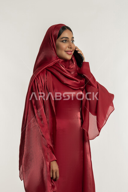 التواصل مع العائلة والأصدقاء, استخدام الأجهزة التقنية الحديثة في الحياة اليومية, مشاركة الأخبار والأحاديث الجيدة, بورتريه لامرأة عربية خليجية إماراتية محجبة ترتدي العباءة الحمراء تمسك بيدها الهاتف المحمول, شابة سعودية تقوم بإجراء اتصال هاتفي, خلفية بيضاء