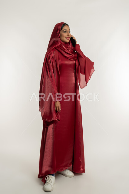 استخدام الأجهزة التقنية الحديثة, التواصل مع العائلة والأصدقاء, مشاركة الأخبار والأحاديث الجيدة, بورتريه لامرأة عربية خليجية إماراتية محجبة ترتدي العباءة الحمراء تمسك بيدها الهاتف المحمول, شابة سعودية تقوم بإجراء اتصال هاتفي, الجسم بالطول الكامل, خلفية بيضاء