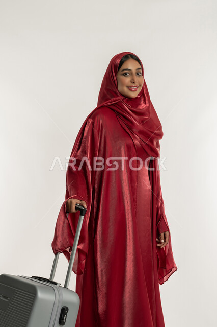 التحضير للإجازة الصيفية, الاستمتاع بالرحلات السياحية في وقت العطلة, التجهيز للسفرات الممتعة, بورتريه لامرأة عربية خليجية إماراتية محجبة ترتدي العباءة الحمراء تمسك بيدها حقيبة سفر رمادية, شابة سعودية تنظر إلى الكاميرا بإيماءات السعادة والسرور, خلفية بيضاء