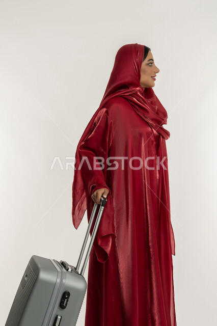 الاستمتاع بالرحلات السياحية في وقت العطلة, الاستعداد لقضاء الأوقات الممتعة, التجهيز للسفرات الممتعة, بورتريه من الجانب لامرأة عربية خليجية إماراتية محجبة ترتدي العباءة الحمراء تمسك بيدها حقيبة سفر رمادية, شابة سعودية تقوم بالتحضير للإجازة الصيفية, خلفية بيضاء