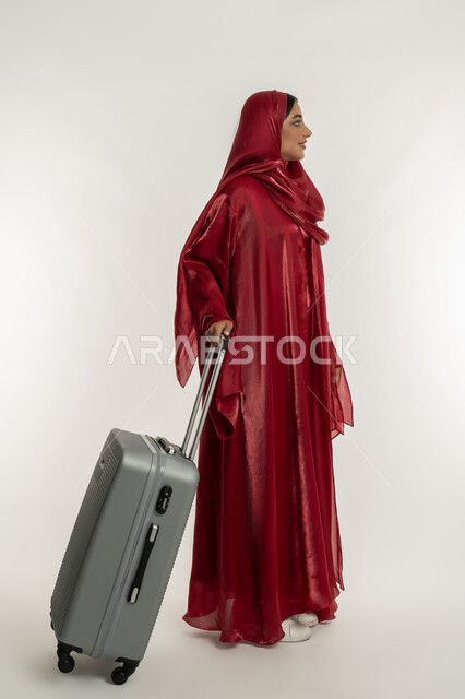 الاستعداد لقضاء الأوقات الممتعة, الاستمتاع بالرحلات السياحية في وقت العطلة، بورتريه لامرأة عربية خليجية إماراتية محجبة ترتدي العباءة الحمراء تمسك بيدها حقيبة سفر رمادية, شابة سعودية تقوم بالتحضير للإجازة الصيفية, الجسم بالطول الكامل, خلفية بيضاء