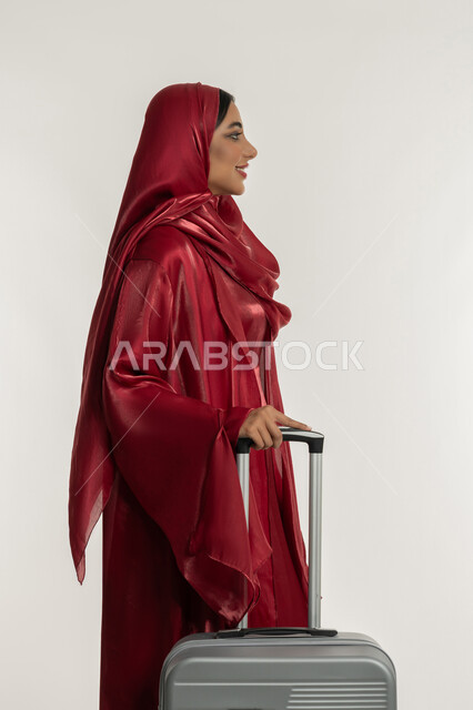 Enjoying vacation trips, preparing for fun times, preparing for fun trips, side portrait of a veiled Emirati Gulf Arab woman wearing a red abaya holding a gray suitcase, a young Saudi woman preparing for summer vacation, white background