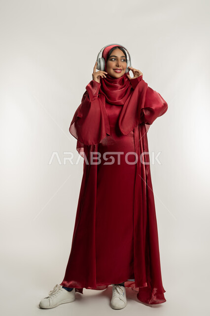 الاستمتاع بسماع الأغاني المفضلة, الاندماج و التفاعل مع الأنغام الموسيقية, استخدام تقنية الهيدفون الحديثة, بورتريه لامرأة عربية خليجية إماراتية محجبة ترتدي عباءة حمراء تضع سماعات الرأس الصوتية, شابة سعودية تستمع إلى الموسيقا, الجسم بالطول الكامل, خلفية بيضاء