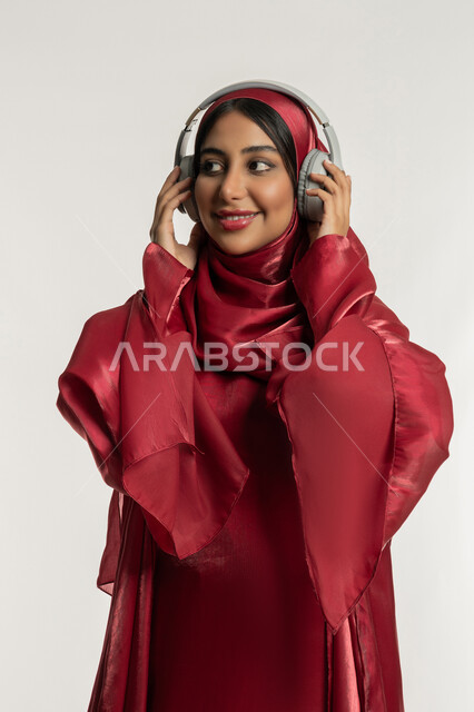 الاندماج و التفاعل مع الأنغام الموسيقية, استخدام تقنية الهيدفون الحديثة, الاستمتاع بسماع الأغاني المفضلة, بورتريه لامرأة عربية خليجية إماراتية محجبة ترتدي العباءة الحمراء تضع سماعات الرأس الصوتية, شابة سعودية تستمع إلى الموسيقا, خلفية بيضاء