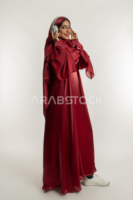 النظر إلى الكاميرا بإيماءات السعادة والسرور, الاندماج مع الأنغام الموسيقية, الاستمتاع بسماع الأغاني, بورتريه لامرأة عربية خليجية إماراتية محجبة ترتدي عباءة حمراء تضع سماعات الرأس الصوتية, شابة سعودية تستمع إلى الموسيقا, الجسم بالطول الكامل, خلفية بيضاء