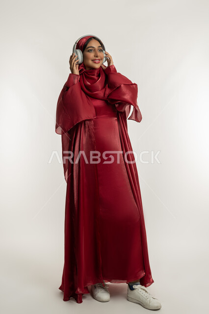 Enjoy listening to your favorite songs, merging and interacting with the musical tunes, using modern headphone technology, portrait of a veiled Emirati Gulf Arab woman wearing a red abaya and wearing audio headphones, a young Saudi woman listening to music, full body, white background
