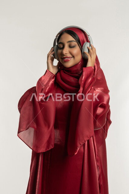 تعابير الاندماج و التفاعل مع الأنغام الموسيقية, استخدام تقنية الهيدفون الحديثة, الاستمتاع بسماع الأغاني المفضلة, بورتريه لامرأة عربية خليجية إماراتية محجبة ترتدي العباءة الحمراء تضع سماعات الرأس الصوتية, شابة سعودية تستمع إلى الموسيقا, خلفية بيضاء