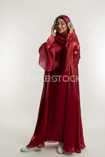Enjoy listening to your favorite songs, merging and interacting with the musical tunes, using modern headphone technology, portrait of a veiled Emirati Gulf Arab woman wearing a red abaya and wearing audio headphones, a young Saudi woman listening to music, full body, white background
