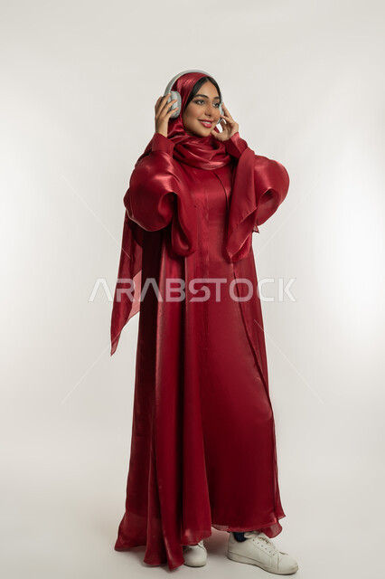 الاندماج و التفاعل مع الأنغام الموسيقية, الاستمتاع بسماع الأغاني المفضلة, استخدام تقنية الهيدفون الحديثة, بورتريه لامرأة عربية خليجية إماراتية محجبة ترتدي عباءة حمراء تضع سماعات الرأس الصوتية, شابة سعودية تستمع إلى الموسيقا, الجسم بالطول الكامل, خلفية بيضاء