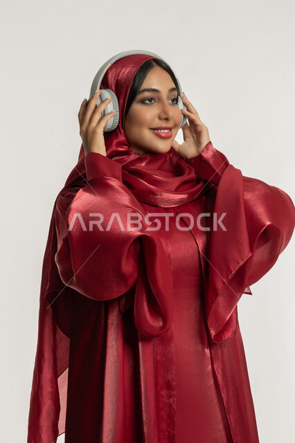 استخدام تقنية الهيدفون الحديثة, الاندماج و التفاعل مع الأنغام الموسيقية, الاستمتاع بسماع الأغاني المفضلة, بورتريه لامرأة عربية خليجية إماراتية محجبة ترتدي العباءة الحمراء تضع سماعات الرأس الصوتية, شابة سعودية تستمع إلى الموسيقا, خلفية بيضاء