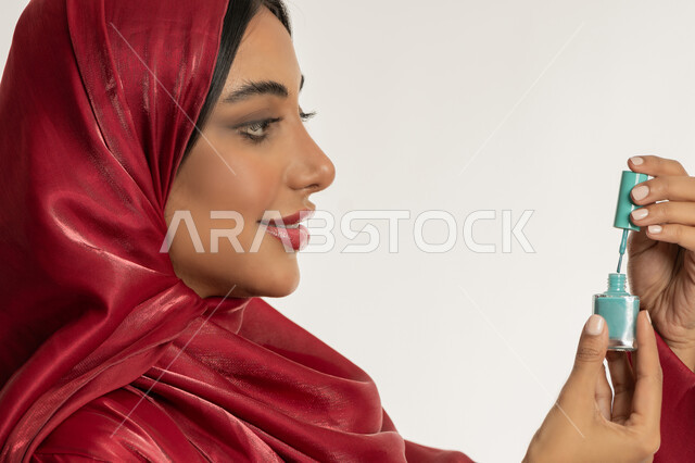 الاهتمام بالأناقة والمظهر الخارجي, مفهوم العناية بالأظافر, التسويق لمنتجات ومستحضرات التجميل, بورتريه مقرب من الجانب لامرأة عربية خليجية إماراتية محجبة ترتدي العباءة الحمراء تمسك بيدها علبة طلاء الأظافر, شابة سعودية تقوم بالتزين والتجمل, خلفية بيضاء