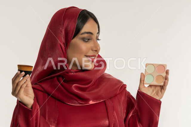 استخدام المساحيق التجميلية, التسويق لمستحضرات التجميل, الاهتمام والعناية بالمظهر الخارجي, مفهوم التزين والتجمل, بورتريه مقرب لامرأة عربية خليجية إماراتية محجبة ترتدي العباءة الحمراء تمسك بيديها فرشاة و علبة أحمر الخدود, شابة سعودية تضع المكياج, خلفية بيضاء