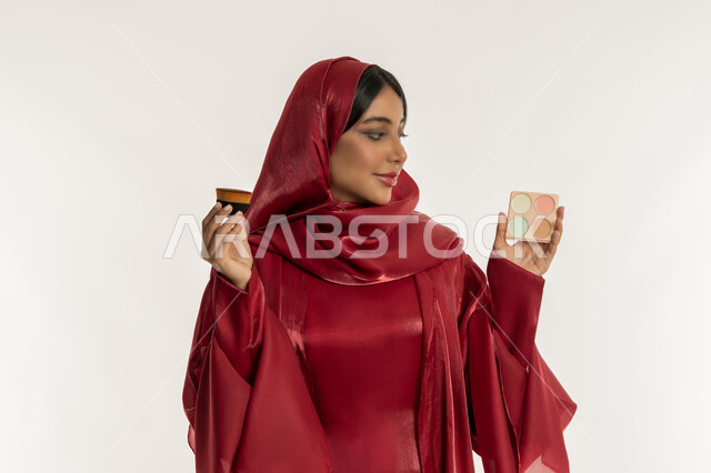 التسويق لمستحضرات التجميل, استخدام المساحيق التجميلية, الاهتمام والعناية بالمظهر الخارجي, مفهوم التزين والتجمل, بورتريه مقرب لامرأة عربية خليجية إماراتية محجبة ترتدي العباءة الحمراء تمسك بيديها فرشاة و علبة أحمر الخدود, شابة سعودية تضع المكياج, خلفية بيضاء