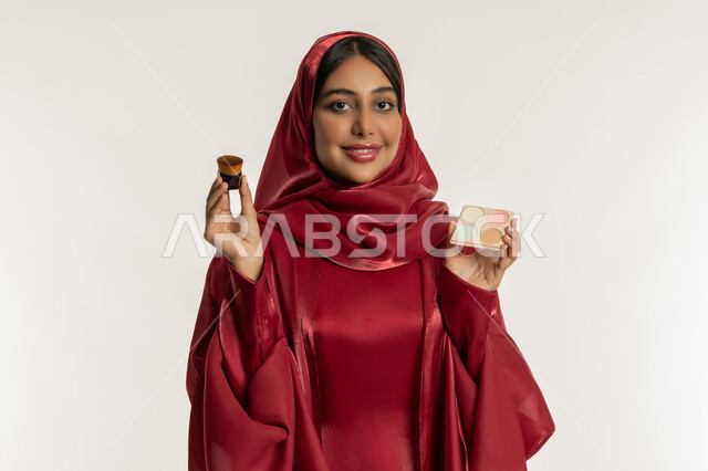مفهوم التزين والتجمل, الشغف بوضع المكياج, الاهتمام والعناية بالبشرة والجمال, بورتريه مقرب لامرأة عربية خليجية إماراتية محجبة ترتدي العباءة الحمراء تمسك بيديها علبة وفرشاة أحمر الخدود, شابة سعودية تنظر إلى الكاميرا بإيماءات السعادة والسرور, خلفية بيضاء
