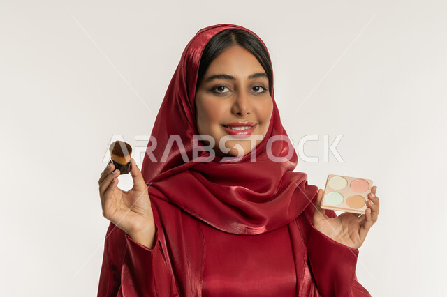 مفهوم التزين والتجمل, الاهتمام والعناية بالبشرة والجمال, الشغف بوضع المكياج, بورتريه مقرب لامرأة عربية خليجية إماراتية محجبة ترتدي العباءة الحمراء تمسك بيديها علبة وفرشاة أحمر الخدود, شابة سعودية تنظر إلى الكاميرا بإيماءات السعادة والسرور, خلفية بيضاء