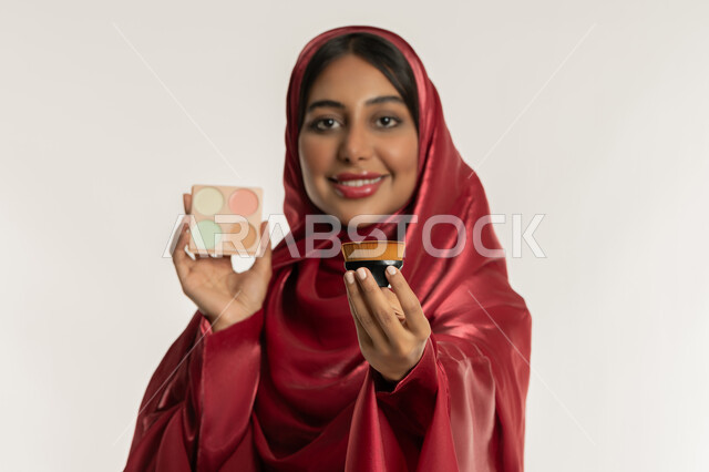 Passion for applying makeup, concept of beautification and beautification, care and attention to skin and beauty, close-up portrait of a veiled Emirati Gulf Arab woman wearing a red abaya holding a blush box and brush in her hands, a young Saudi woman looking at the camera with gestures of happiness and pleasure, white background