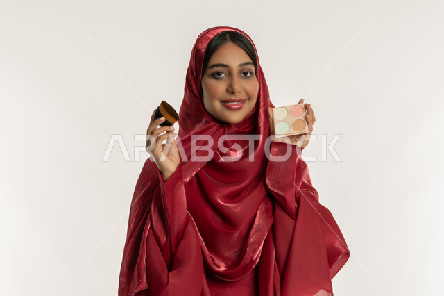 الاهتمام والعناية بالبشرة والجمال, مفهوم التزين والتجمل, الشغف بوضع المكياج, بورتريه مقرب لامرأة عربية خليجية إماراتية محجبة ترتدي العباءة الحمراء تمسك بيديها علبة وفرشاة أحمر الخدود, شابة سعودية تنظر إلى الكاميرا بإيماءات السعادة والسرور, خلفية بيضاء