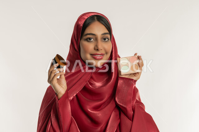 مفهوم التزين والتجمل, الاهتمام والعناية بالبشرة والجمال, الشغف بوضع المكياج, بورتريه مقرب لامرأة عربية خليجية إماراتية محجبة ترتدي العباءة الحمراء تمسك بيديها علبة وفرشاة أحمر الخدود, شابة سعودية تنظر إلى الكاميرا بإيماءات السعادة والسرور, خلفية بيضاء