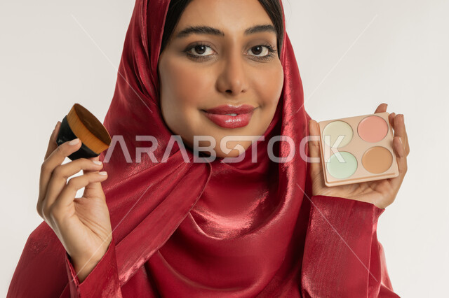 الشغف بوضع المكياج, مفهوم التزين والتجمل, الاهتمام والعناية بالبشرة والجمال, بورتريه مقرب لامرأة عربية خليجية إماراتية محجبة ترتدي العباءة الحمراء تمسك بيديها علبة وفرشاة أحمر الخدود, شابة سعودية تنظر إلى الكاميرا بإيماءات السعادة والسرور, خلفية بيضاء