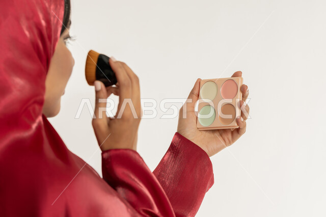 التسويق لمستحضرات التجميل, استخدام المساحيق التجميلية, الاهتمام بالمظهر الخارجي, مفهوم التزين والتجمل, بورتريه مقرب من الخلف لامرأة عربية خليجية إماراتية محجبة ترتدي العباءة الحمراء تستعمل علبة وفرشاة أحمر الخدود, شابة سعودية تضع المكياج, خلفية بيضاء