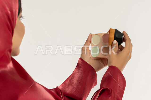مفهوم التزين والتجمل, الاهتمام بالمظهر الخارجي, استخدام المساحيق التجميلية, التسويق لمستحضرات التجميل, بورتريه مقرب من الخلف لامرأة عربية خليجية إماراتية محجبة ترتدي العباءة الحمراء تستعمل علبة وفرشاة أحمر الخدود, شابة سعودية تضع المكياج, خلفية بيضاء