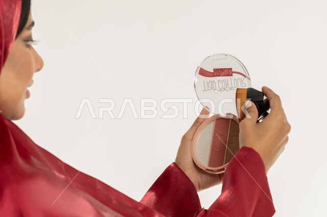 التسويق لمستحضرات التجميل, استخدام المساحيق التجميلية, الاهتمام بالمظهر الخارجي, مفهوم التزين والتجمل, بورتريه مقرب من الخلف لامرأة عربية خليجية إماراتية محجبة ترتدي العباءة الحمراء تستعمل علبة وفرشاة أحمر الخدود, شابة سعودية تضع المكياج, خلفية بيضاء