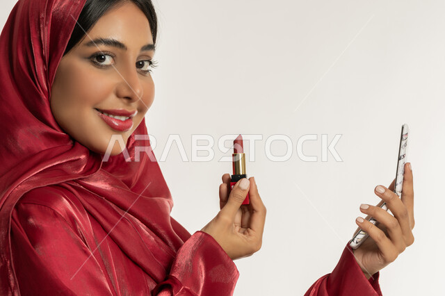 العناية بالجمال والمظهر الخارجي, الترويج لمنتجات و مساحيق التجميل, استعمال المستحضرات التجميلية, مفهوم التزين والتجمل, بورتريه مقرب من الجانب لامرأة عربية خليجية إماراتية محجبة ترتدي العباءة الحمراء تضع أحمر شفاه, شابة سعودية تقوم بوضع المكياج, خلفية بيضاء