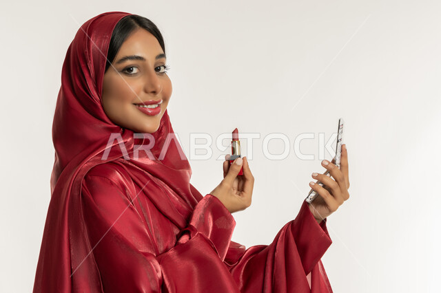 مفهوم التزين والتجمل, استعمال المستحضرات التجميلية, الترويج لمنتجات و مساحيق التجميل, العناية بالجمال والمظهر الخارجي, بورتريه مقرب من الجانب لامرأة عربية خليجية إماراتية محجبة ترتدي العباءة الحمراء تضع أحمر شفاه, شابة سعودية تقوم بوضع المكياج, خلفية بيضاء