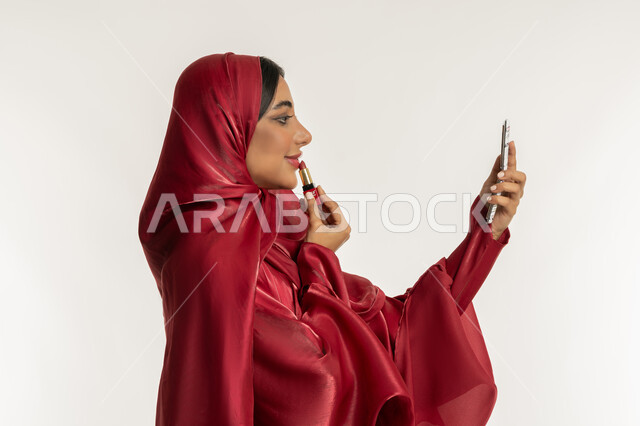 العناية بالجمال والمظهر الخارجي, الترويج لمنتجات و مساحيق التجميل, استعمال المستحضرات التجميلية, مفهوم التزين والتجمل, بورتريه مقرب من الجانب لامرأة عربية خليجية إماراتية محجبة ترتدي العباءة الحمراء تضع أحمر شفاه, شابة سعودية تقوم بوضع المكياج, خلفية بيضاء