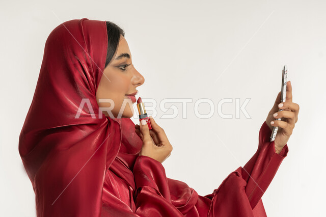 مفهوم التزين والتجمل, استعمال المستحضرات التجميلية, الترويج لمنتجات و مساحيق التجميل, العناية بالجمال والمظهر الخارجي, بورتريه مقرب من الجانب لامرأة عربية خليجية إماراتية محجبة ترتدي العباءة الحمراء تضع أحمر شفاه, شابة سعودية تقوم بوضع المكياج, خلفية بيضاء