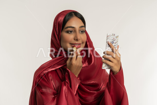 استعمال المستحضرات التجميلية, مفهوم التزين والتجمل, الترويج لمنتجات و مساحيق التجميل, العناية بالجمال والمظهر الخارجي, بورتريه مقرب لامرأة عربية خليجية إماراتية محجبة ترتدي العباءة الحمراء تضع أحمر شفاه, شابة سعودية تقوم بوضع المكياج, خلفية بيضاء