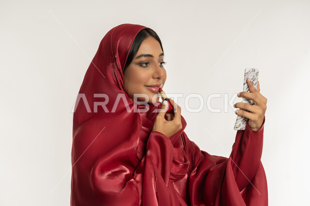 الترويج لمنتجات و مساحيق التجميل, استعمال المستحضرات التجميلية, مفهوم التزين والتجمل, العناية بالجمال والمظهر الخارجي, بورتريه مقرب لامرأة عربية خليجية إماراتية محجبة ترتدي العباءة الحمراء تضع أحمر شفاه, شابة سعودية تقوم بوضع المكياج, خلفية بيضاء